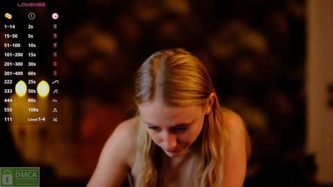 Media: Video of a young blonde woman with straight hair, wearing a strapless top, looking down. Background is blurred with warm lighting.