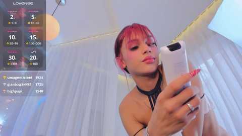 Media: Video of a young woman with light skin and red hair, wearing a black halter top, holding a phone, in a bright, modern room with white curtains.