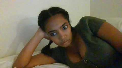 Media: Video of a young Black woman with dark skin, resting her head on her hand, wearing a dark green shirt, on a white bed in a dimly lit room.