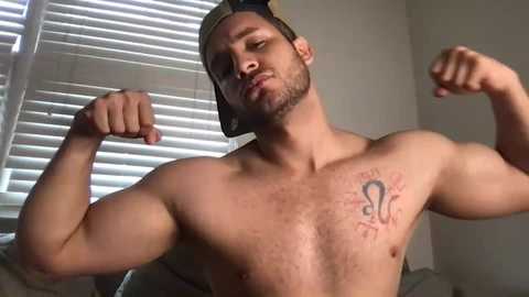 Media: Video of a muscular, shirtless man with a tattoo of a spider on his chest, flexing his biceps. He wears a backwards baseball cap. Background shows closed white blinds.