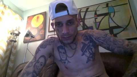 Media: Video of a shirtless tattooed man with a white cap, sitting on a couch in a room with abstract art and a lamp.