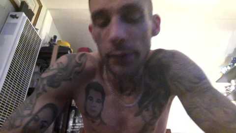 Media: A close-up video of a shirtless, heavily tattooed man with a short buzz cut, smiling, with a white ceiling fan and shelves in the background.