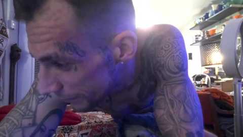 Media: Video of a tattooed man with a beard, blue-tinted, leaning on a bed with patterned bedding, in a dimly lit room with shelves and a lamp.