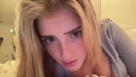 Media: Video of a blonde woman with fair skin, wearing a gray cardigan, leaning forward with a serious expression, in a dimly lit room with a beige wall and white bed in the background.