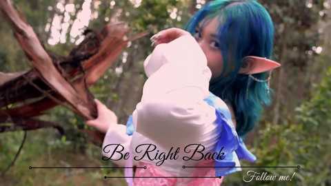 Media: Video of a young woman with bright blue hair and elf ears, wearing a white and pink cosplay outfit, holding a white hat, in a forest setting.