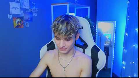 Media: Video of a young, shirtless man with curly blonde hair in a gaming chair, wearing a gold necklace, in a blue-lit room with posters, a plant, and a gaming setup in the background.