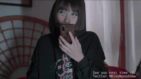 Media: Video of a woman with dark bob haircut, pale skin, wearing a black hoodie with a colorful graphic print, holding a phone to her face, standing in a room with a red wooden chair and white curtain.