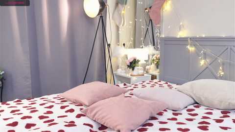 Media: Video of a romantic, pastel-toned bedroom with a white bedspread adorned with red heart patterns, two pink heart-shaped pillows, and fairy lights.