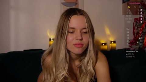 Media: Video of a topless blonde woman with long, wavy hair, seated on a dark couch, background featuring candles and a partially visible Christmas tree.