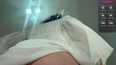 Media: Video of a person with dark hair, wearing a white hospital gown, lying on a hospital bed with a bright blue surgical light overhead. A digital display in the upper right corner shows vital signs.