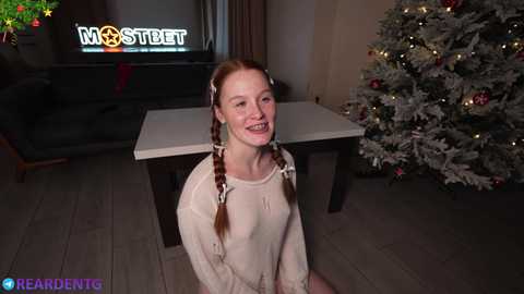 Media: A video of a smiling, fair-skinned woman with red pigtails, wearing a beige sweater, kneeling in front of a Christmas tree in a cozy living room.