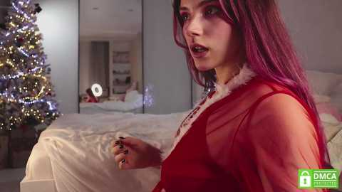 Media: A video of a young woman with long, purple hair, wearing a sheer, red, Santa-themed outfit, standing in a modern, dimly-lit bedroom with a Christmas tree and white bed.