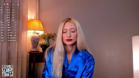 Video of a blonde woman with fair skin, red lips, and closed eyes, wearing a blue satin robe, sitting in a softly lit room with a lamp and potted plant.