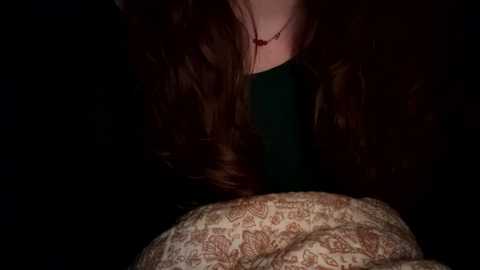 Media: Video of a person with long brown hair, wearing a dark green top and a patterned blanket, in a dimly lit room.