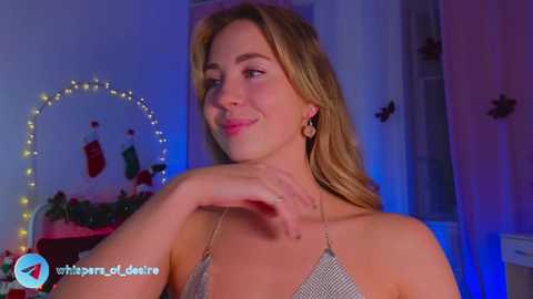 Media: Video of a smiling, fair-skinned woman with long, blonde hair, wearing a silver sequined halter top, standing in a dimly lit room with Christmas decorations and fairy lights.