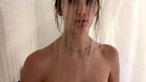 Media: Video of a topless woman with wet hair, wearing a silver necklace, standing in a white-tiled shower. She has a neutral expression, with water droplets visible on her skin.