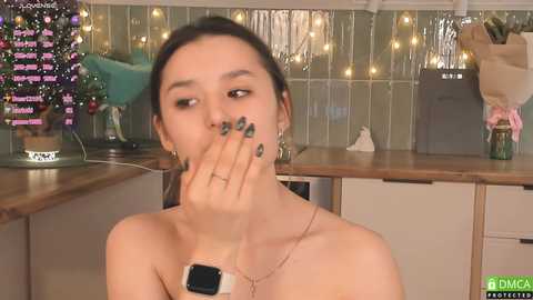 Media: Video of a woman with dark hair and green nail polish, wearing a smartwatch, covering her mouth in a kitchen with festive decorations.