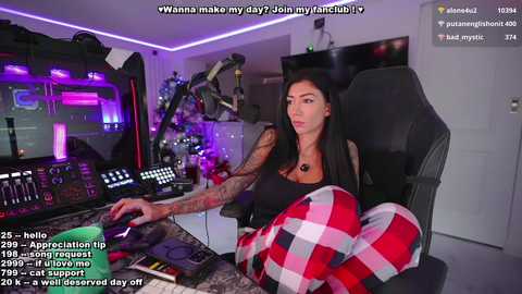 Media: Video of a tattooed woman with long black hair, wearing a black top, playing video games in a modern gaming room with purple lighting.