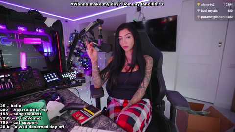 Media: A video of a tattooed woman in a black tank top and red plaid skirt, sitting at a DJ booth with various music equipment. She has long black hair and a serious expression.