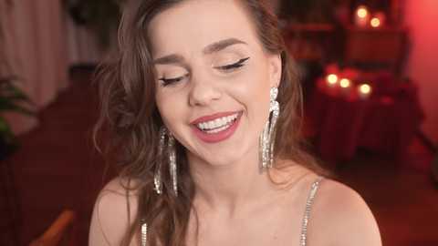 Media: Video of a smiling woman with long brown hair, wearing sparkling earrings and a sleeveless dress, in a dimly lit, warmly decorated room with red candles.