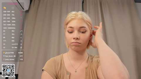 Media: Video of a fair-skinned, blonde woman with short hair, wearing a beige top, adjusting her hair in a mirror. Background features grey curtains and a digital overlay showing social media metrics.