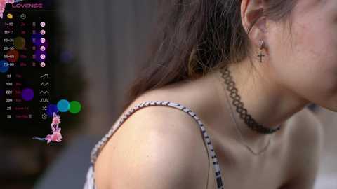 Media: Video of a young woman with light skin, dark hair, wearing a black choker and a black and white patterned top. Background shows a blurred, dimly lit room with colorful chat bubbles.