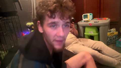 Media: Video of a man with curly hair, wearing a striped jacket, holding a baby, sitting in a cluttered kitchen with toys and a green step stool.