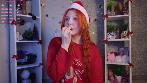 Video of a fair-skinned woman with long red hair, wearing a Santa hat and red sweater, eating a cookie in a festive, decorated room with shelves adorned with toys and Christmas ornaments.