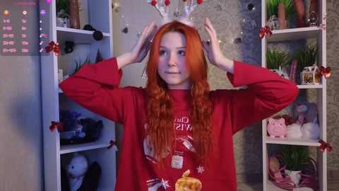 Video of a young woman with long red hair, wearing a red Christmas sweater and a white Santa hat, adjusting it, in a festive, cozy room with shelves filled with holiday decorations.