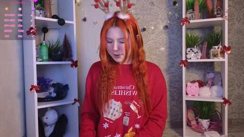 Video of a fair-skinned woman with long, vibrant orange hair, wearing a red Santa Claus sweater, standing in a festive room with white shelves adorned with Christmas decorations.