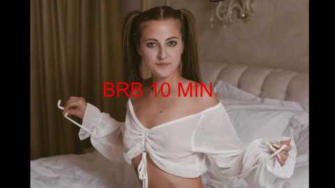 Media: Video of a fair-skinned woman with long brown hair styled in pigtails, wearing a white off-shoulder crop top, standing in a bedroom with a white bed and beige curtains.