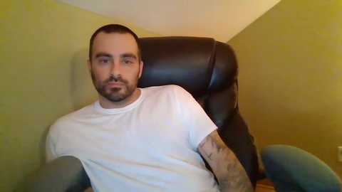 Media: Video of a bearded man with a shaved head, wearing a white t-shirt and gray pants, seated in a black leather chair against a mustard-yellow wall.