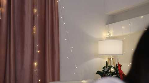 Media: Video of a cozy bedroom corner with pink curtains, white walls, and fairy lights; a table lamp with a white shade and red and green Christmas decorations, creating a festive, warm atmosphere.