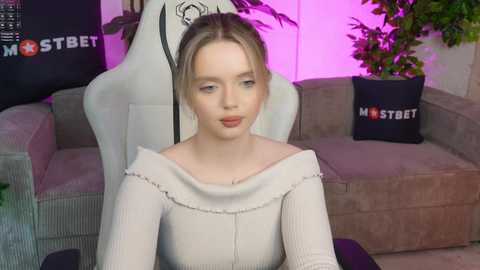 Media: Video of a young woman with fair skin and blonde hair in a white ribbed off-the-shoulder top, sitting in a white gaming chair, surrounded by plants, a couch, and a \"MOSTBET\" pillow.