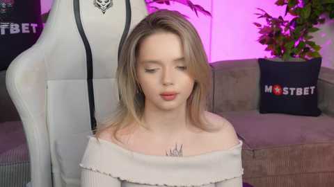 Media: Video of a fair-skinned, blonde woman in a white off-shoulder sweater, with a tattoo on her chest, standing in a modern living room with a gaming chair and a plant.