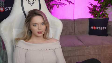 Media: Video of a young Caucasian woman with light skin, blonde hair, and blue eyes, wearing a white off-shoulder sweater, sitting in a white gaming chair. Background features a gray couch with a black pillow saying \"Masterson\" and green plants.