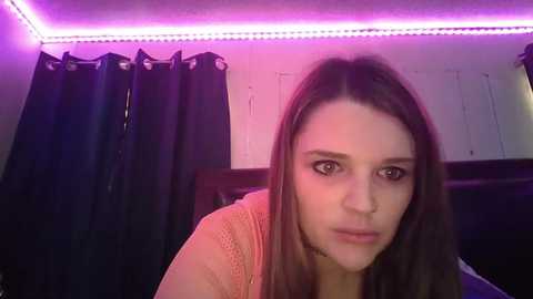 Video of a young woman with straight, brown hair, wearing a beige sweater, indoors under purple LED strip lights. Dark curtains and white walls in the background.