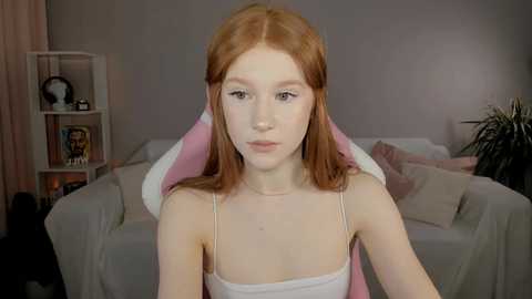 Media: A young woman with long, straight red hair and fair skin, wearing a white spaghetti-strap top, sits on a pink and white chair in a cozy, dimly lit living room with a white couch and beige curtains.