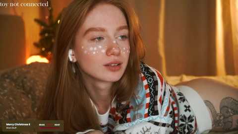 Media: Video of a fair-skinned, freckled woman with long red hair, wearing a colorful Christmas sweater, lying on a bed, looking into the camera, surrounded by warm, cozy indoor lighting.