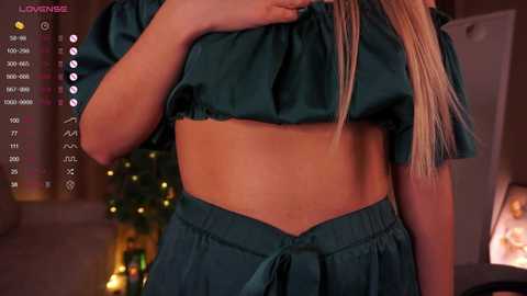 Media: Video of a woman lifting her teal satin crop top, revealing her toned midriff. Background shows a dimly lit room with a Christmas tree and string lights.