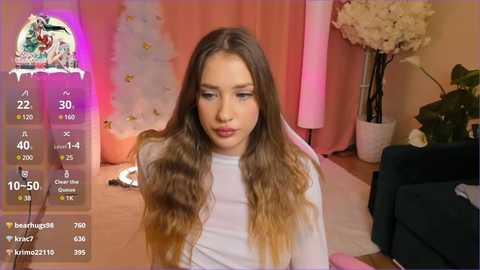 Media: Video of a young woman with long, wavy brown hair, wearing a white top, in a pink room with floral decor and a green plant.