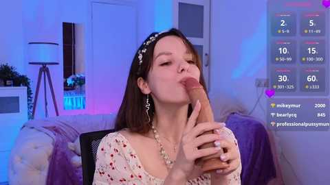 Media: Video of a fair-skinned woman with long brown hair, wearing a white floral dress, kneeling and performing oral sex on a large, erect penis, in a modern bedroom with purple and blue lighting, social media stats overlay.