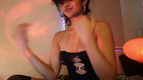 Media: Video of a young woman with short, dark hair, wearing a black, strapless, cutout dress, standing in a dimly lit room with a leopard-print blanket and orange lamp.