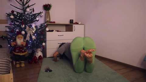 Media: Video of a person in green bodysuit on all fours beside a decorated Christmas tree and wooden dresser in a cozy, dimly-lit room.