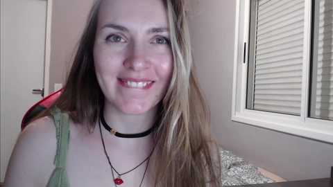 Media: Video of a smiling, fair-skinned woman with long, straight blonde hair, wearing a green tank top, black choker, and two necklaces. Background includes white blinds and beige walls.