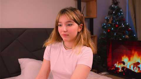 Media: Video of a young woman with light skin and blonde hair, wearing a white t-shirt, sitting on a bed with a grey headboard, next to a lit Christmas tree and fireplace.