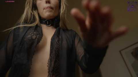 Media: Video of a topless blonde woman with long hair, wearing a sheer black lace top, black collar, and raised hand, set against a dimly lit, blurred background.