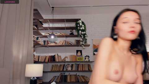 Video of a topless woman with long black hair, small breasts, and fair skin, standing in a modern, minimalist room with white walls, wooden shelves filled with books, and a white lamp.