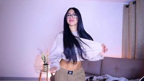 Media: Video of a slender, long-haired Asian woman with glasses, wearing a white crop top and beige shorts, standing in a modern, dimly lit room with a beige couch, a vase, and a window.