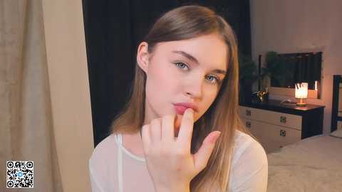 Media: Video of a young Caucasian woman with straight, light brown hair, wearing a white long-sleeved top, seductively biting her finger in a bedroom with a lit lamp and a bed.